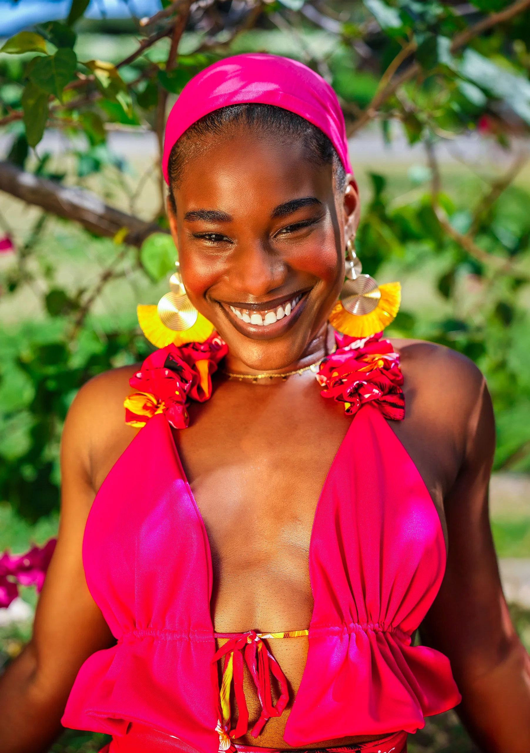 Smiling woman wearing a pink headwrap and yellow earrings among green leaves